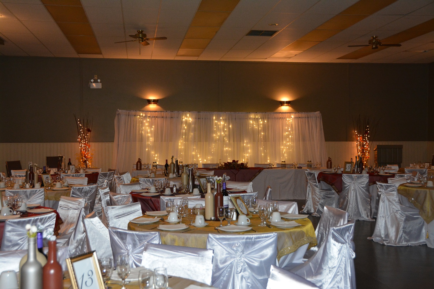 room with tables decorated with yellow tableclothes and set with white dishes, chairs around the tables with white covers, a head table with a backdrop hanging behind with lights